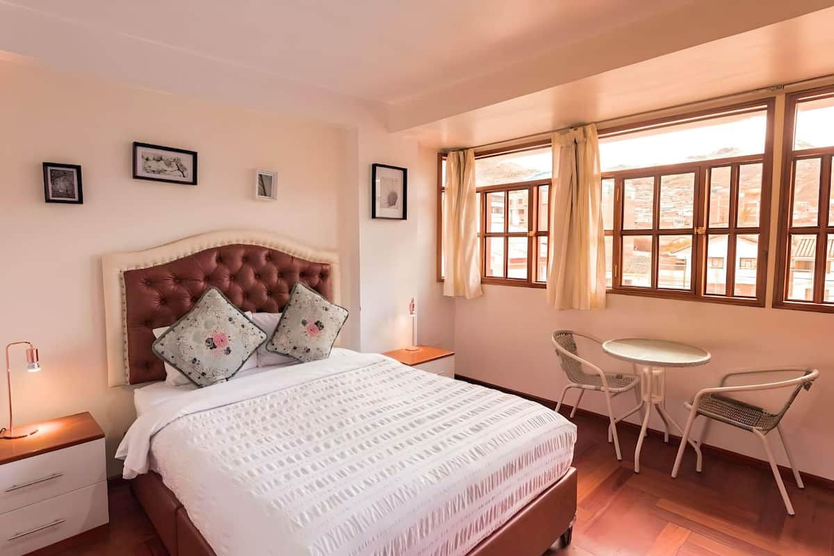 A bedroom with a large bed, a bedside table, a lamp, a chair, a small table, and a window with curtains.
