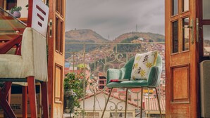 A room with a view of a cityscape, a green chair with a floral pillow, and a wooden chair with a white cushion. - Cozy Room Cusco (Cusco)
