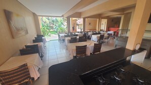 A restaurant with multiple tables and chairs, a kitchen area with a stove, and a view of greenery outside. - Pousada Porto das Dunas (Aquiraz)