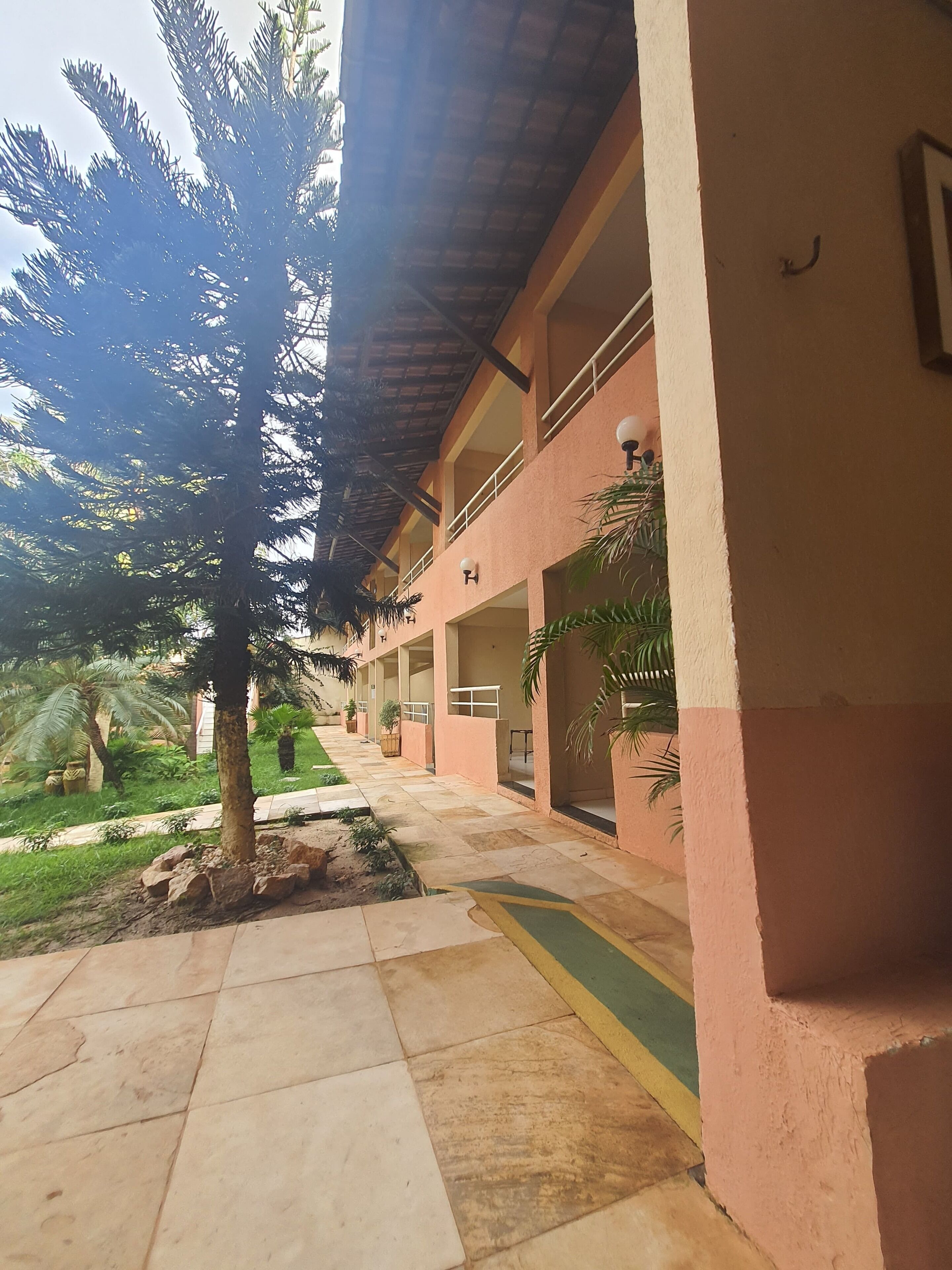 A covered outdoor area with a tiled walkway, palm trees, and a building with balconies.