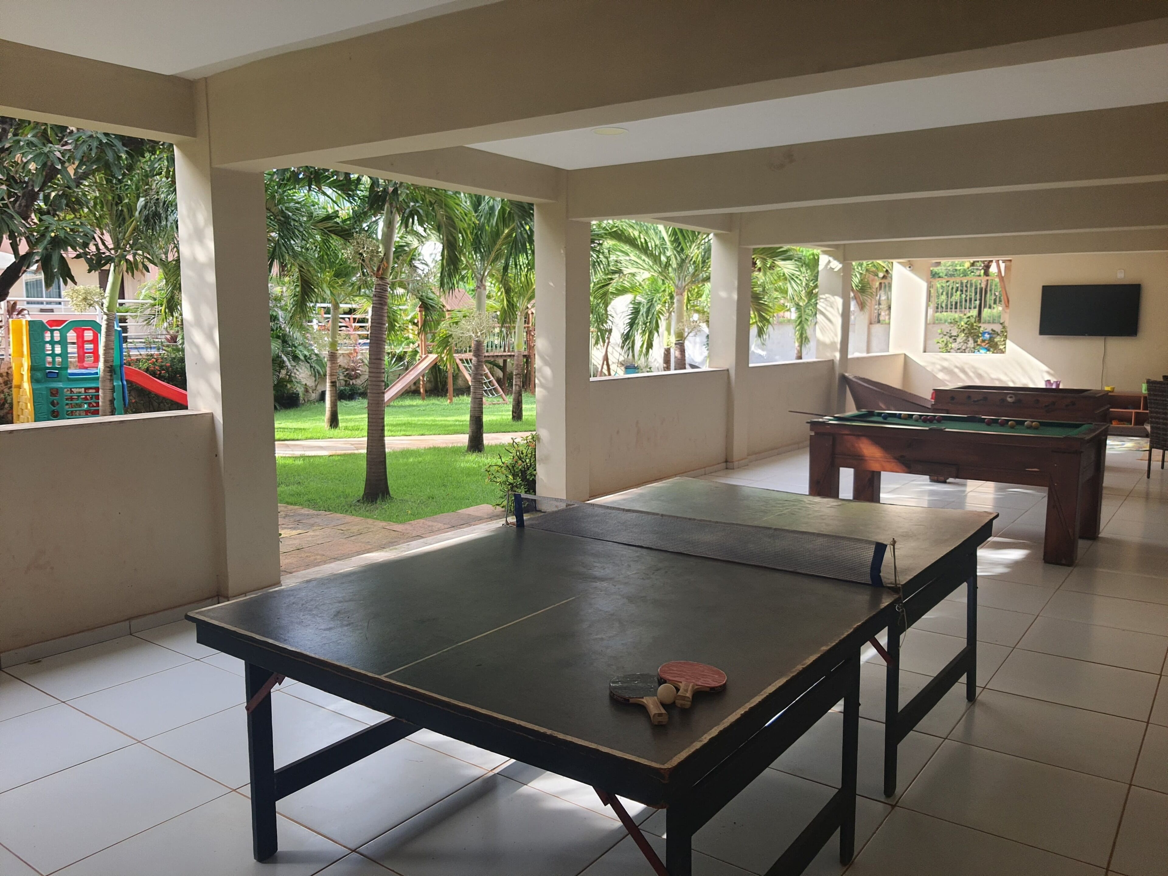 A spacious indoor area with a ping pong table, a pool table, and a view of a playground with slides and palm trees.