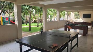 A spacious indoor area with a ping pong table, a pool table, and a view of a playground with slides and palm trees.