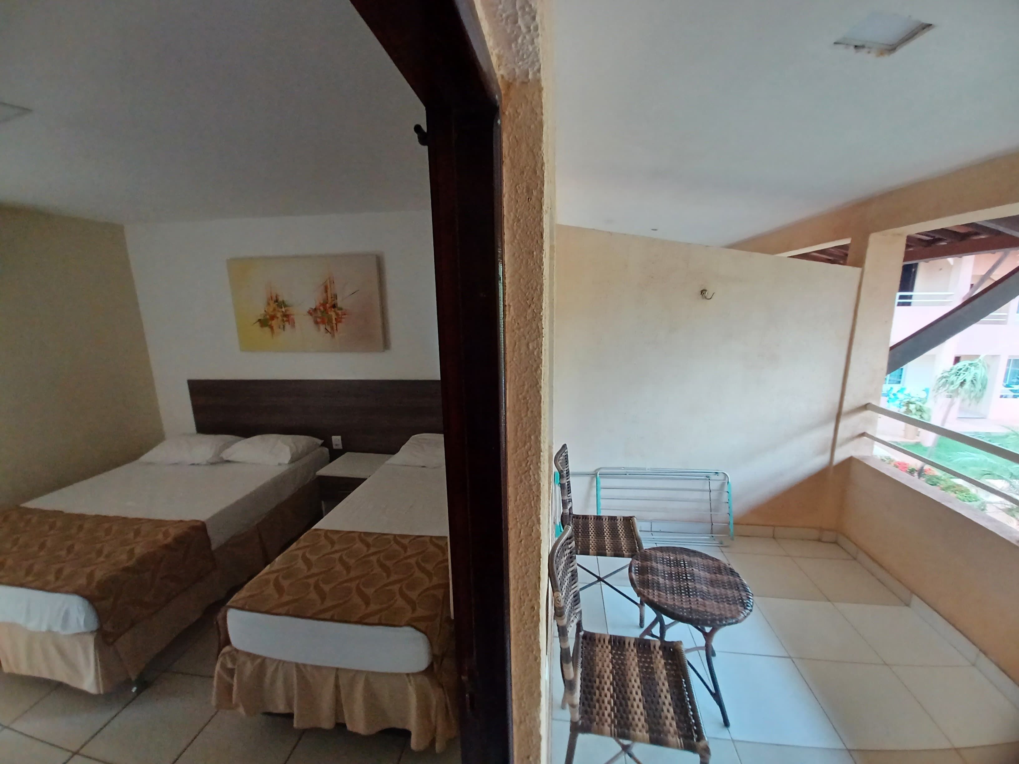 Ein Zimmer mit zwei Einzelbetten, einem kleinen Tisch mit Stühlen, einem an der Wand hängenden Kunstwerk und einem Balkon mit Blick auf Grünflächen.