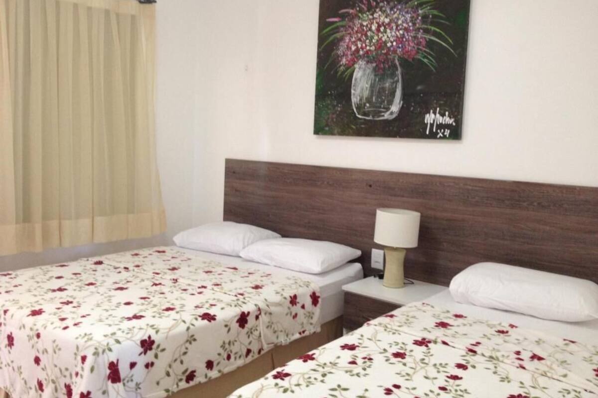 A hotel room with two beds, a wooden headboard, a nightstand with a lamp, and a floral painting on the wall.