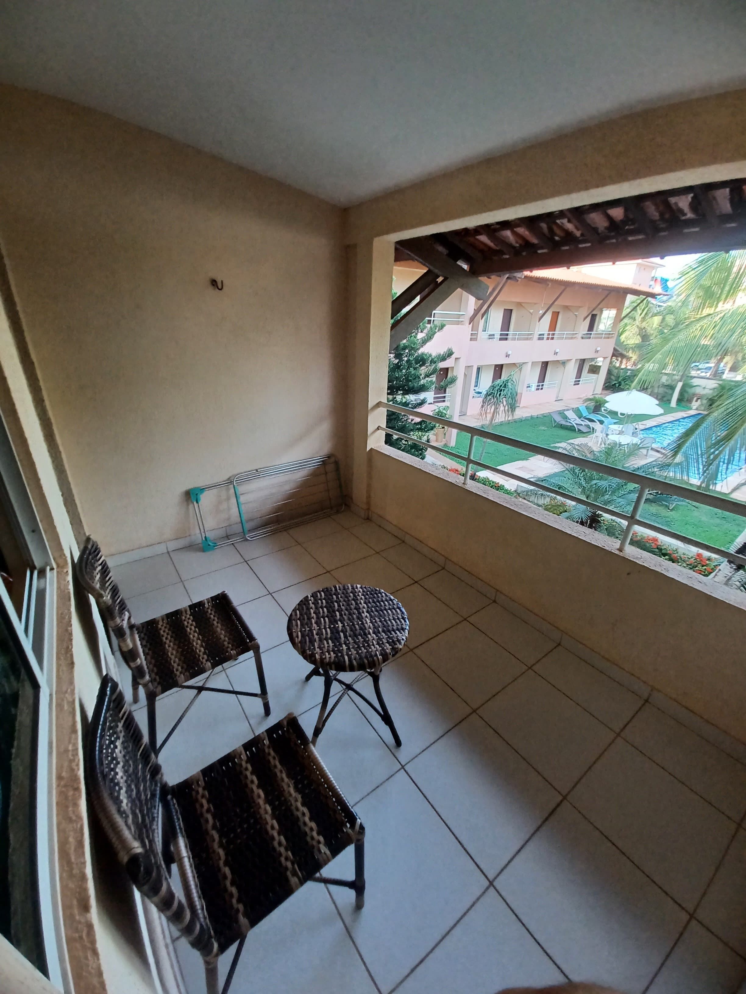 A balcony with two wicker chairs and a small table, a tiled floor, and a view of a building and a swimming pool.