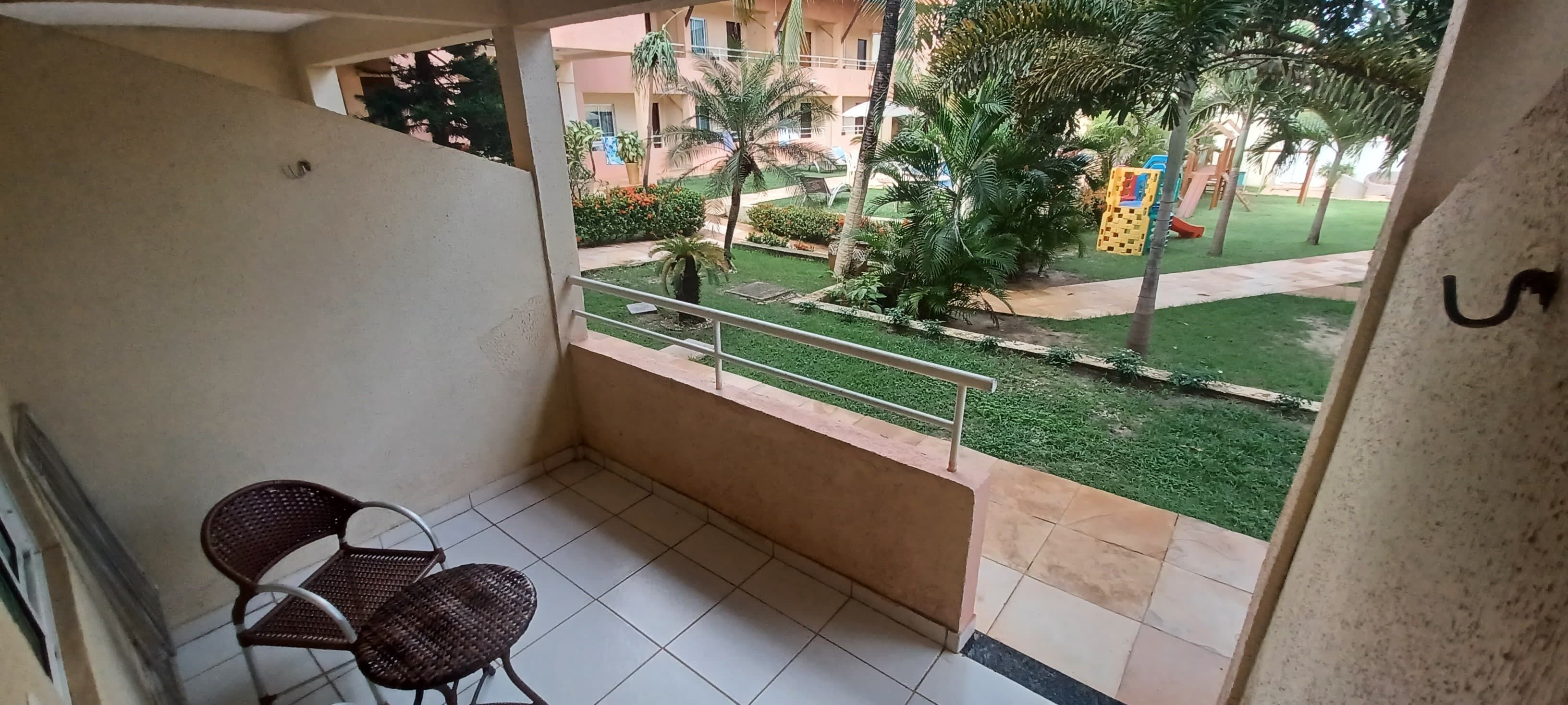 A balcony with two wicker chairs, a tiled floor, and a view of a well-maintained garden with palm trees and a playground.