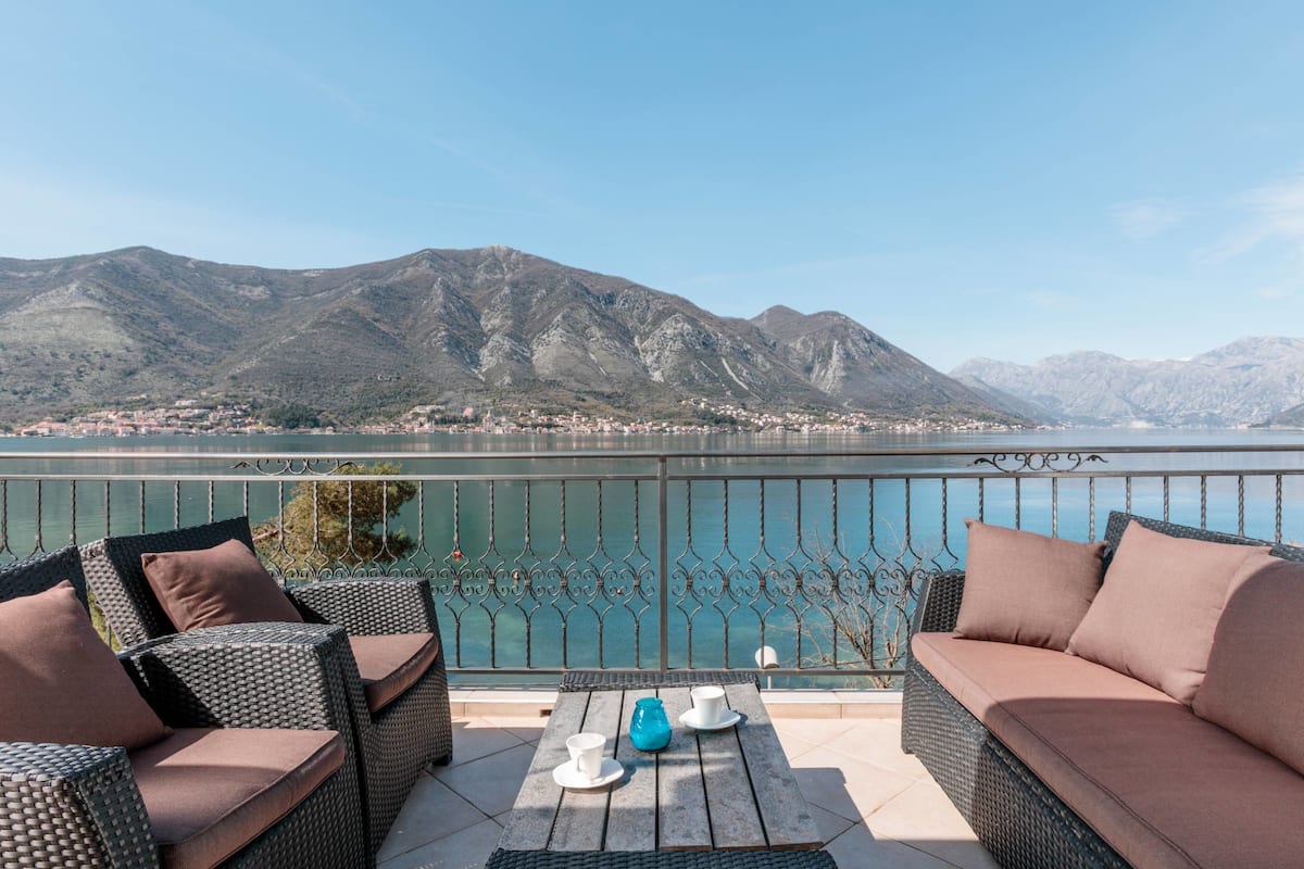 Una terraza con mobiliario de mimbre, una mesa con tazas y un florero, y vistas a montañas y un lago.
