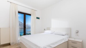 A bedroom with a large bed, a nightstand, a window with curtains, and a view of mountains. - Apartments Miramar (Kotor)