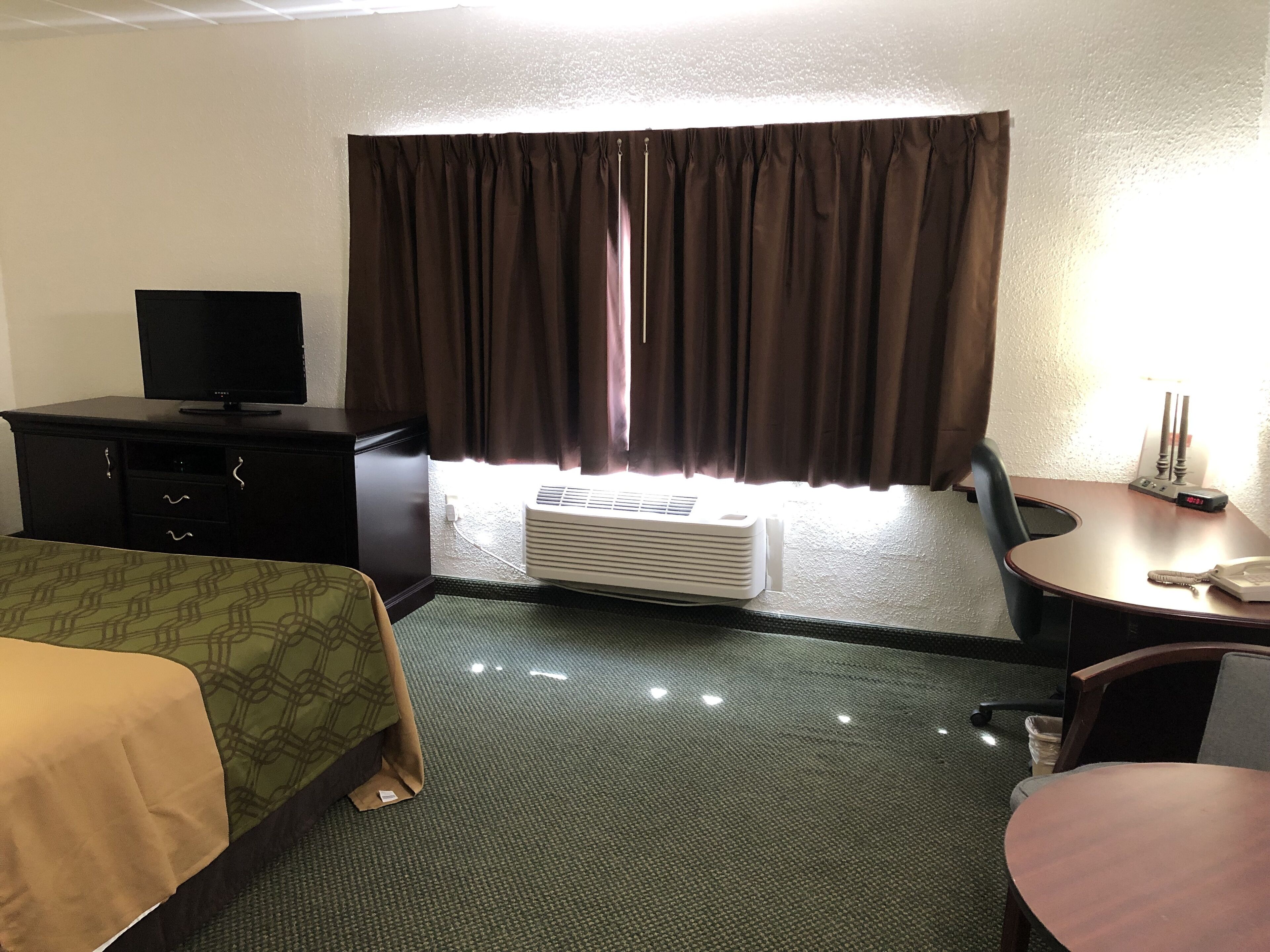 A hotel room with a bed, a desk, a chair, a television, an air conditioning unit, and a window with curtains.
