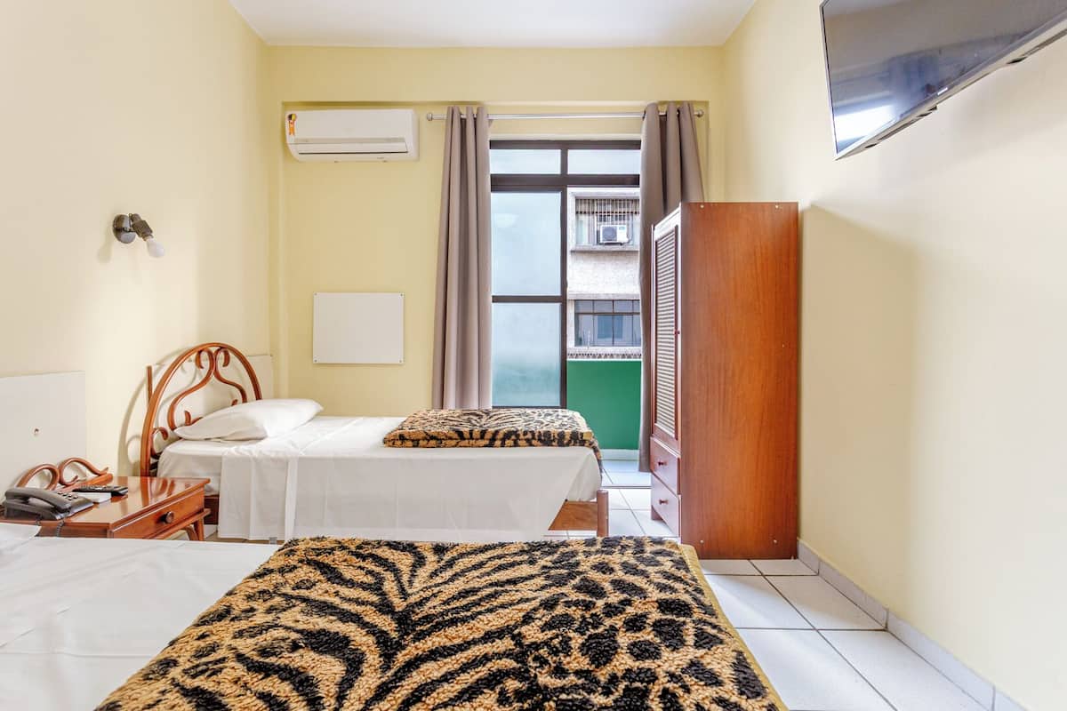A hotel room with two beds, a wooden wardrobe, a desk with a telephone and remote controls, an air conditioning unit, and a view of a building through a window with curtains.