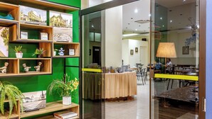 Una hall d'albergo moderna con parete verde, mensole in legno con oggetti decorativi, un divisorio in vetro e un'area ristorazione con tavoli e sedie.