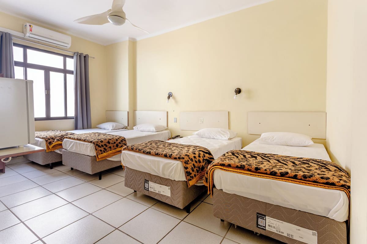 A hotel room with four single beds, an air conditioning unit, a ceiling fan, and a window with curtains.