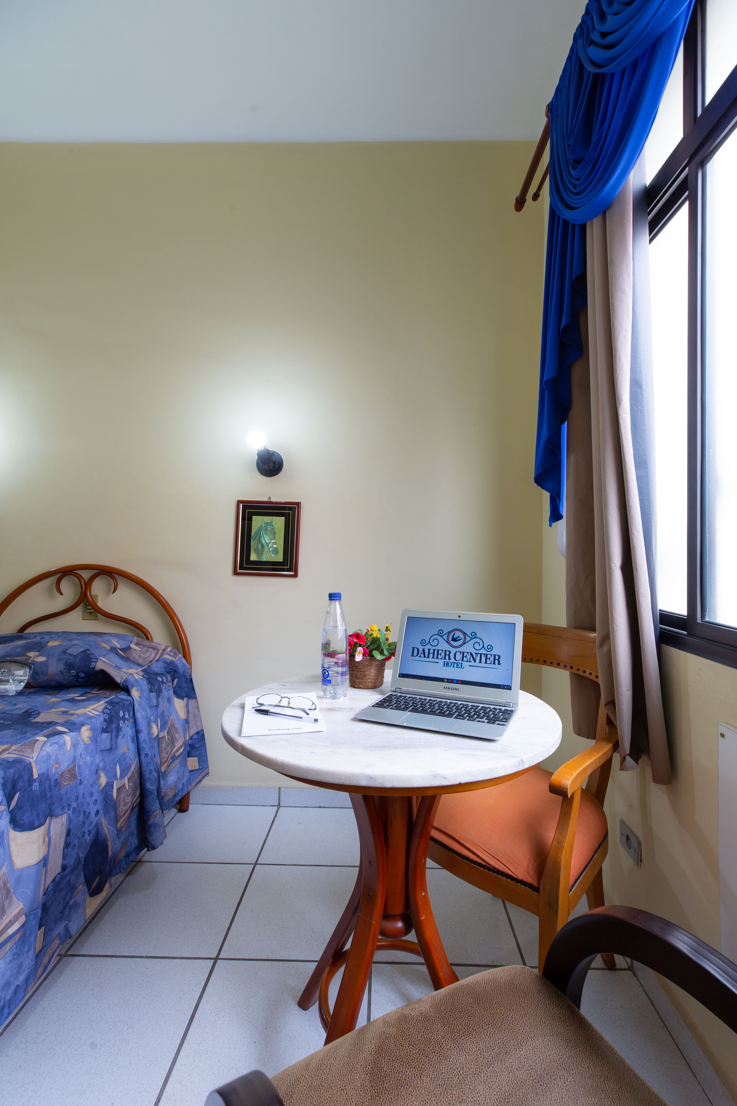 A hotel room with a bed, a desk with a laptop, a chair, a small table, and a window with curtains.