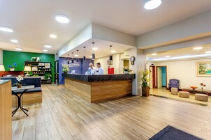 Un espace d’accueil d’hôtel comprenant un comptoir en marbre, un bureau d’accueil, une horloge et un coin salon avec un canapé et des fauteuils.