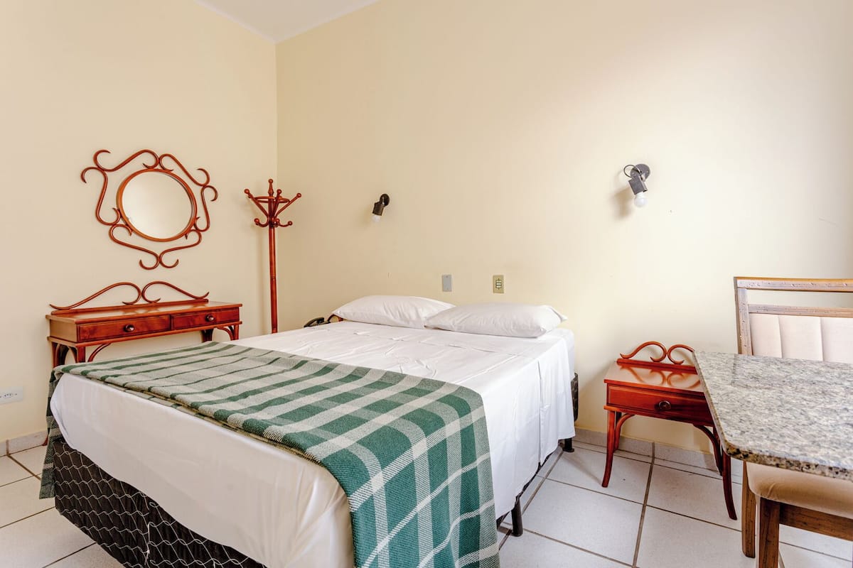 A bedroom with a bed, a wooden nightstand, a chair, a small table, a mirror, and a coat rack.