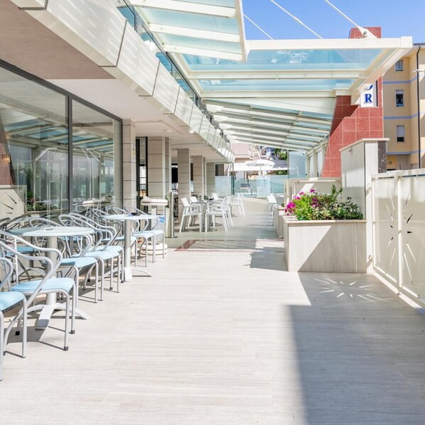 A sunlit outdoor deck with glass railings, tables, and chairs, and a building with a red facade in the background. - Hotel Astor (Alba Adriatica)