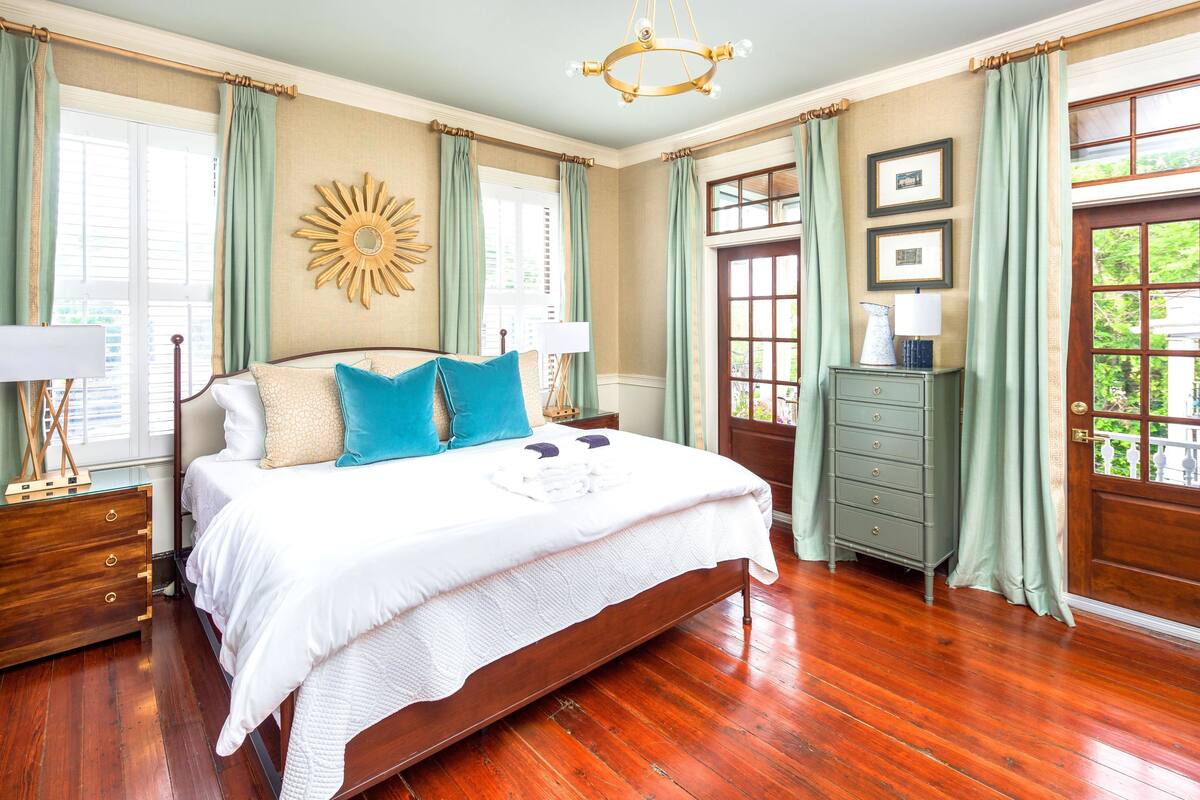 Quarto espaçoso com uma cama grande, piso de madeira, cortinas verdes e um adorno de parede em formato de raio de sol.
