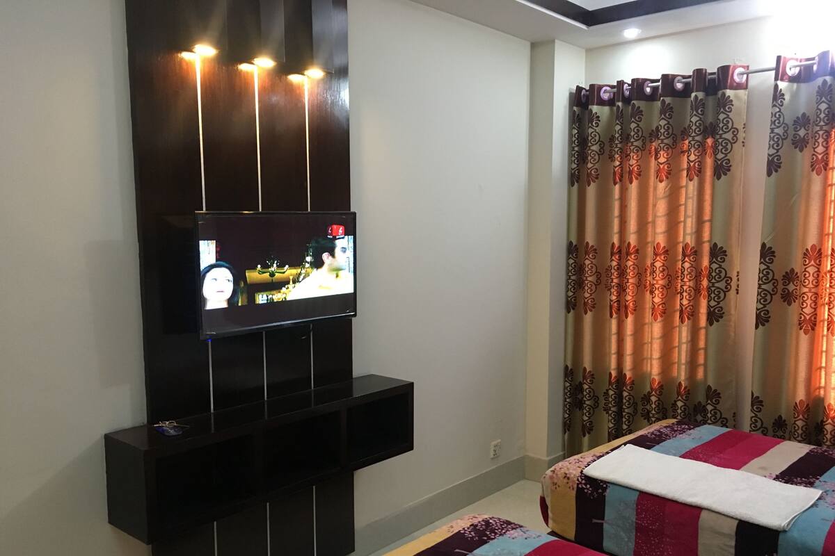 Ein Schlafzimmer mit an der Wand montiertem Fernseher, einem Bett mit farbenfroher Steppdecke und einem Vorhang mit Blumenmuster.