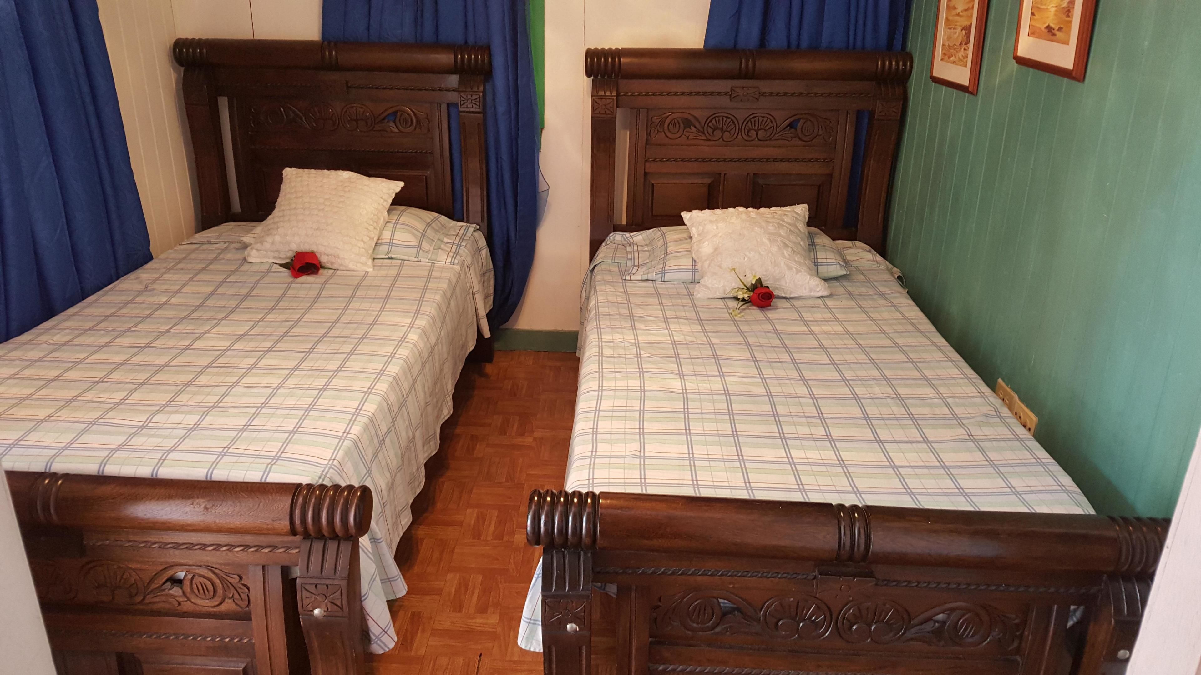 Dos camas de madera con cabeceras, una cortina azul y una pared verde con cuadros enmarcados.