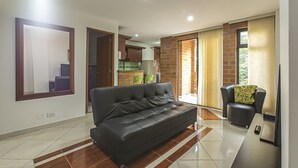 A modern living room with a black leather sofa, a wooden coffee table, and a small armchair with a patterned cushion. The room has a tiled floor and a large mirror on the wall. - Zojo Island (Medellín)