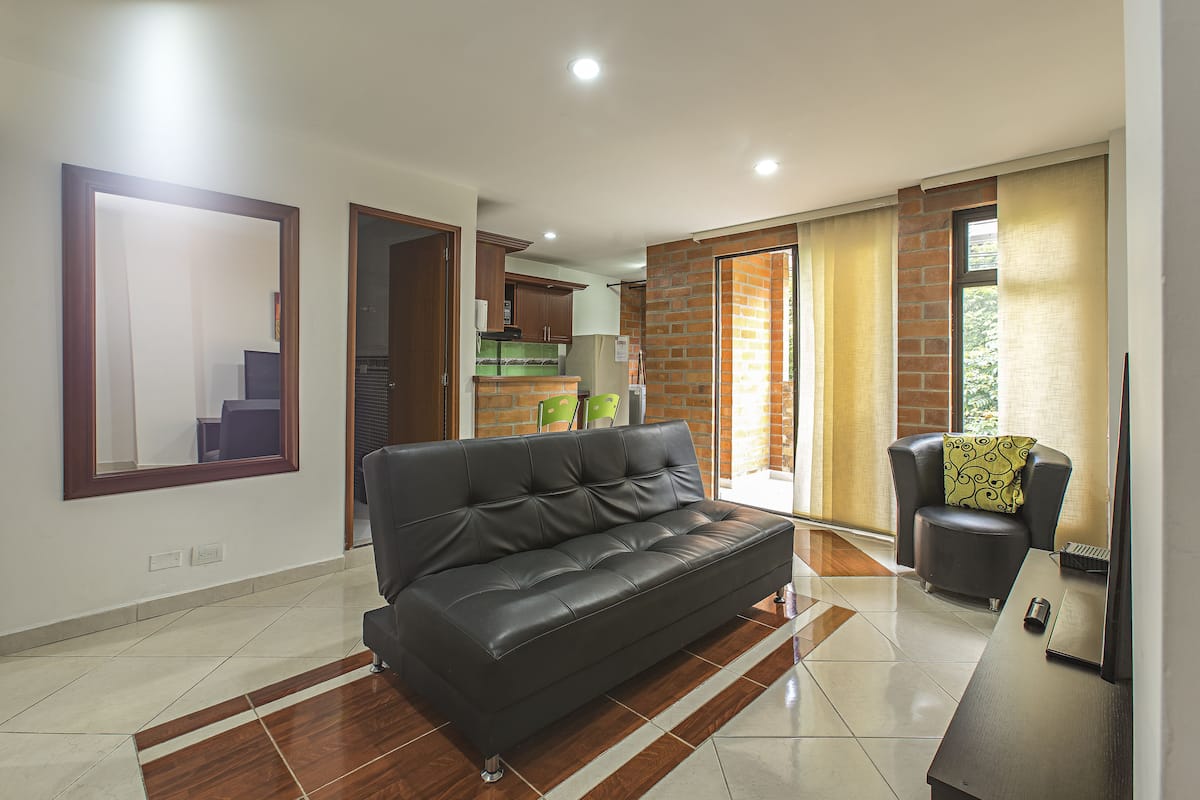 A modern living room with a black leather sofa, a wooden coffee table, and a small armchair with a patterned cushion. The room has a tiled floor and a large mirror on the wall.