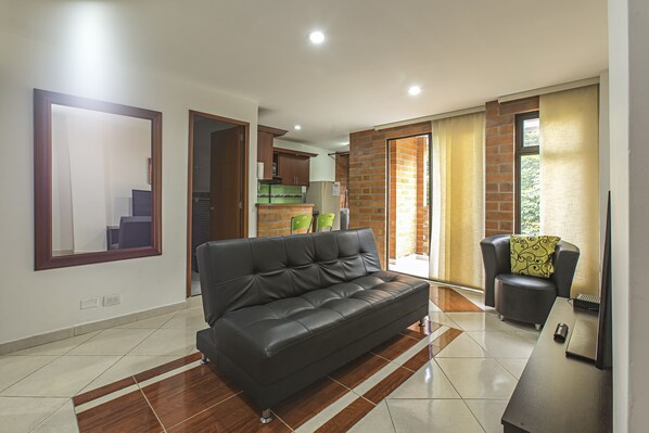 A modern living room with a black leather sofa, a wooden coffee table, and a small armchair with a patterned cushion. The room has a tiled floor and a large mirror on the wall.