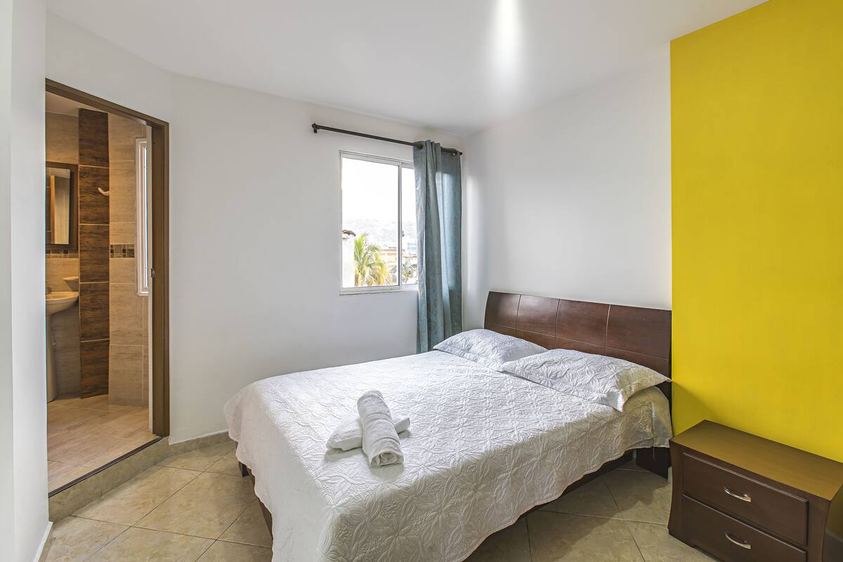 A bedroom with a bed, a window with curtains, a wooden headboard, and a yellow wall.