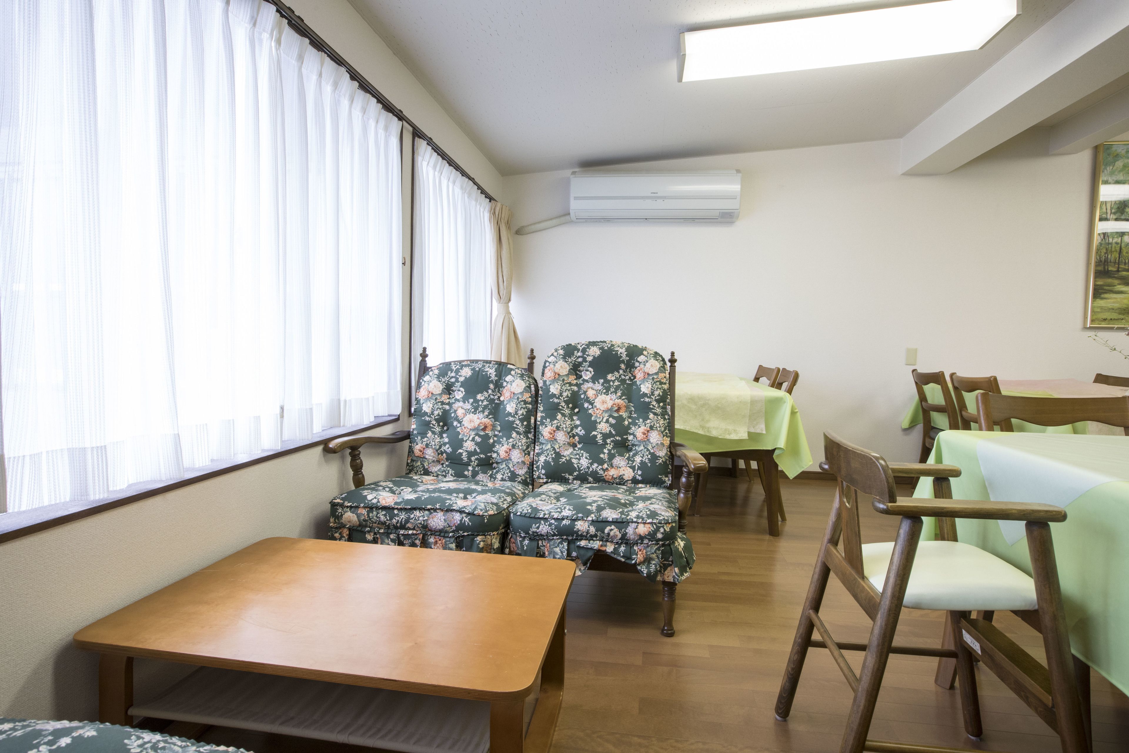 A room with a wooden table, two floral armchairs, a wooden chair, and a dining table with green tablecloths.