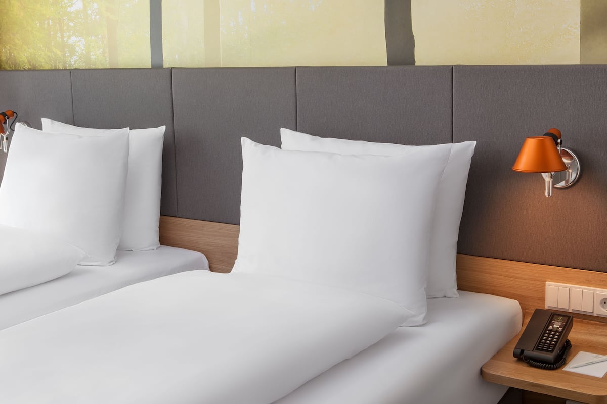 A hotel room with two beds, grey headboard, white bedding, two wall lamps, a bedside table with a phone and notepad, and a power outlet.