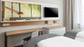A modern hotel room with a large flat-screen TV, a desk with a lamp, a chair, and a bed with grey bedding.