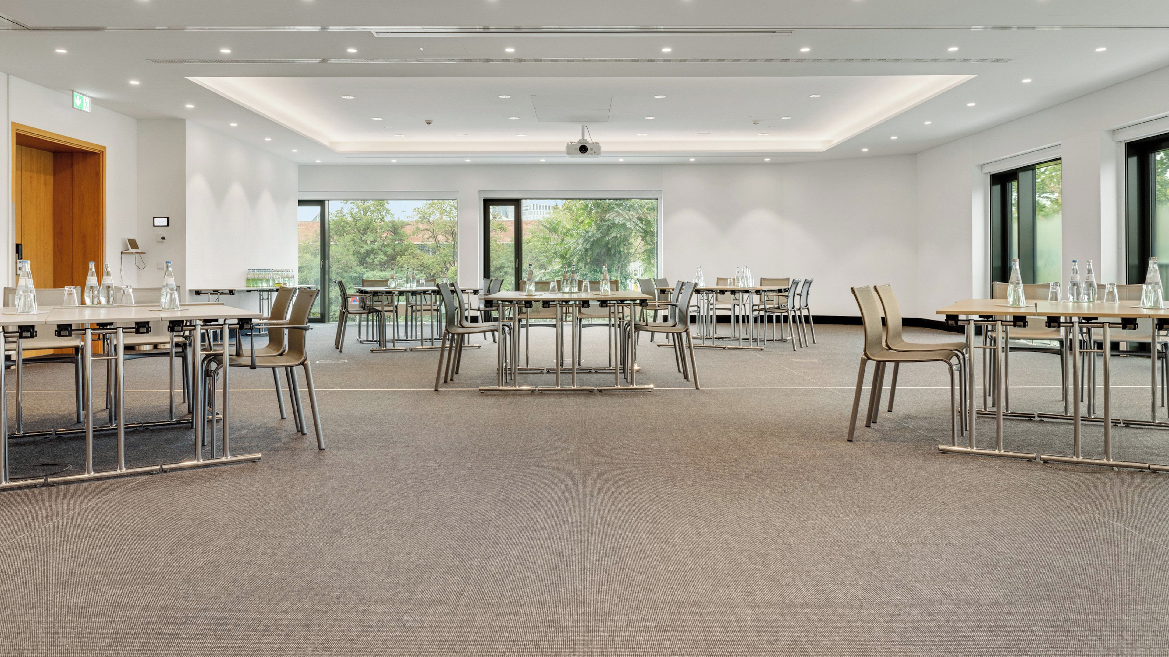 Ein Konferenzraum mit mehreren Tischen und Stühlen, großen Fenstern und einem Teppichboden.