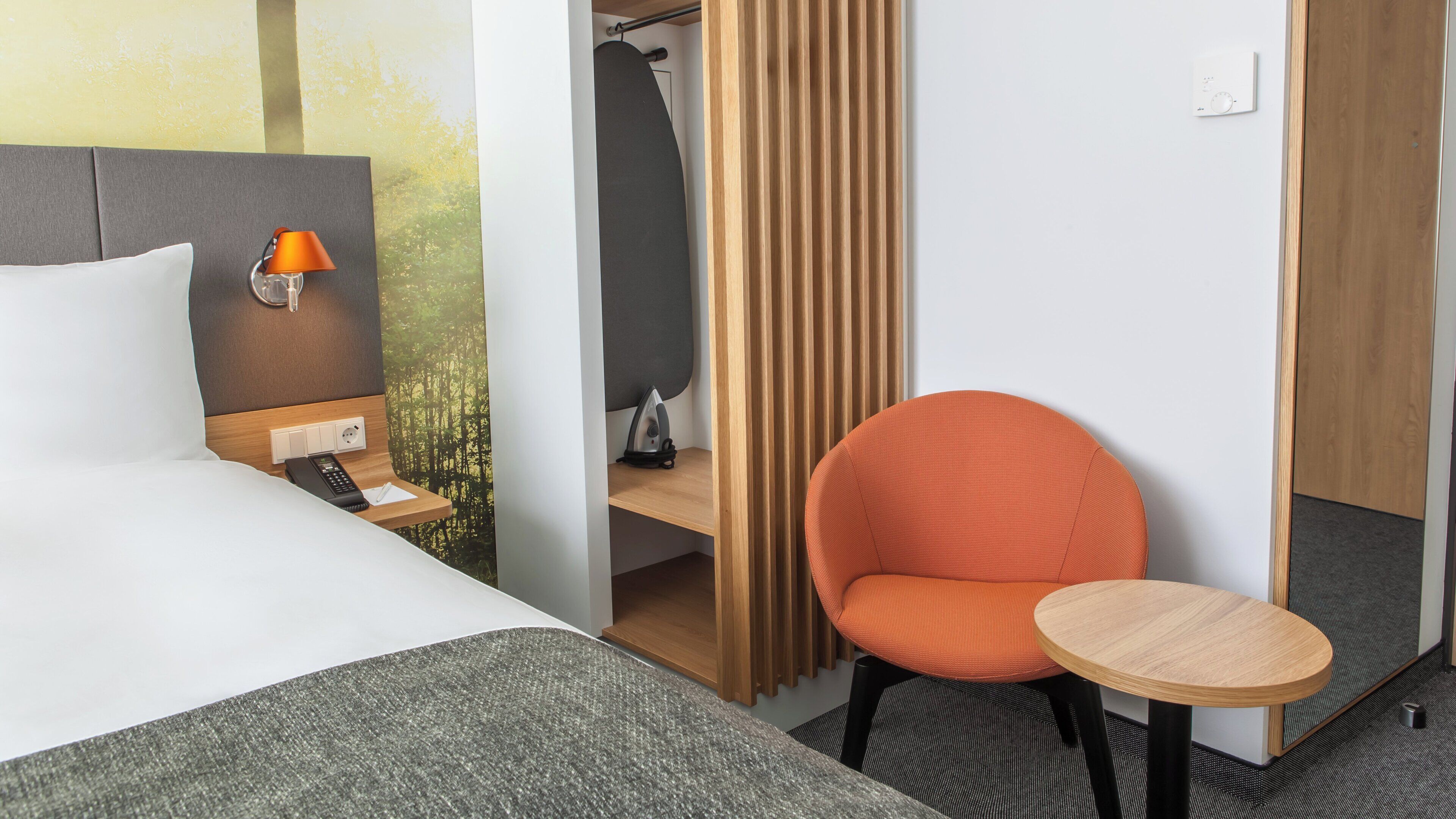 A hotel room with a bed, a desk with a lamp, a chair, and a small table.