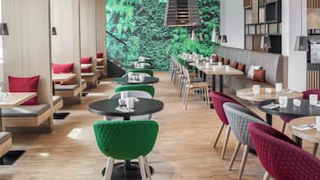 Et moderne restaurantmiljø med runde borde, grønne og røde polstrede stole og et grønt vægmaleri.