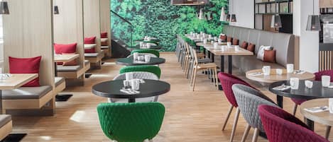Un interior de restaurante moderno con mesas redondas, sillas tapizadas en tonos verde y rojo, y un mural de follaje en la pared.