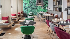 A modern restaurant interior with round tables, green and red upholstered chairs, and a wall mural of greenery. - Holiday Inn Munich - Leuchtenbergring by IHG (Munich)