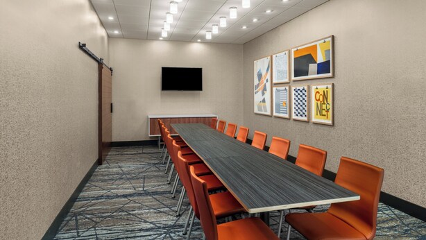 Un salón de conferencias con una larga mesa, sillas naranjas, un televisor de pantalla plana y obras de arte enmarcadas en la pared.