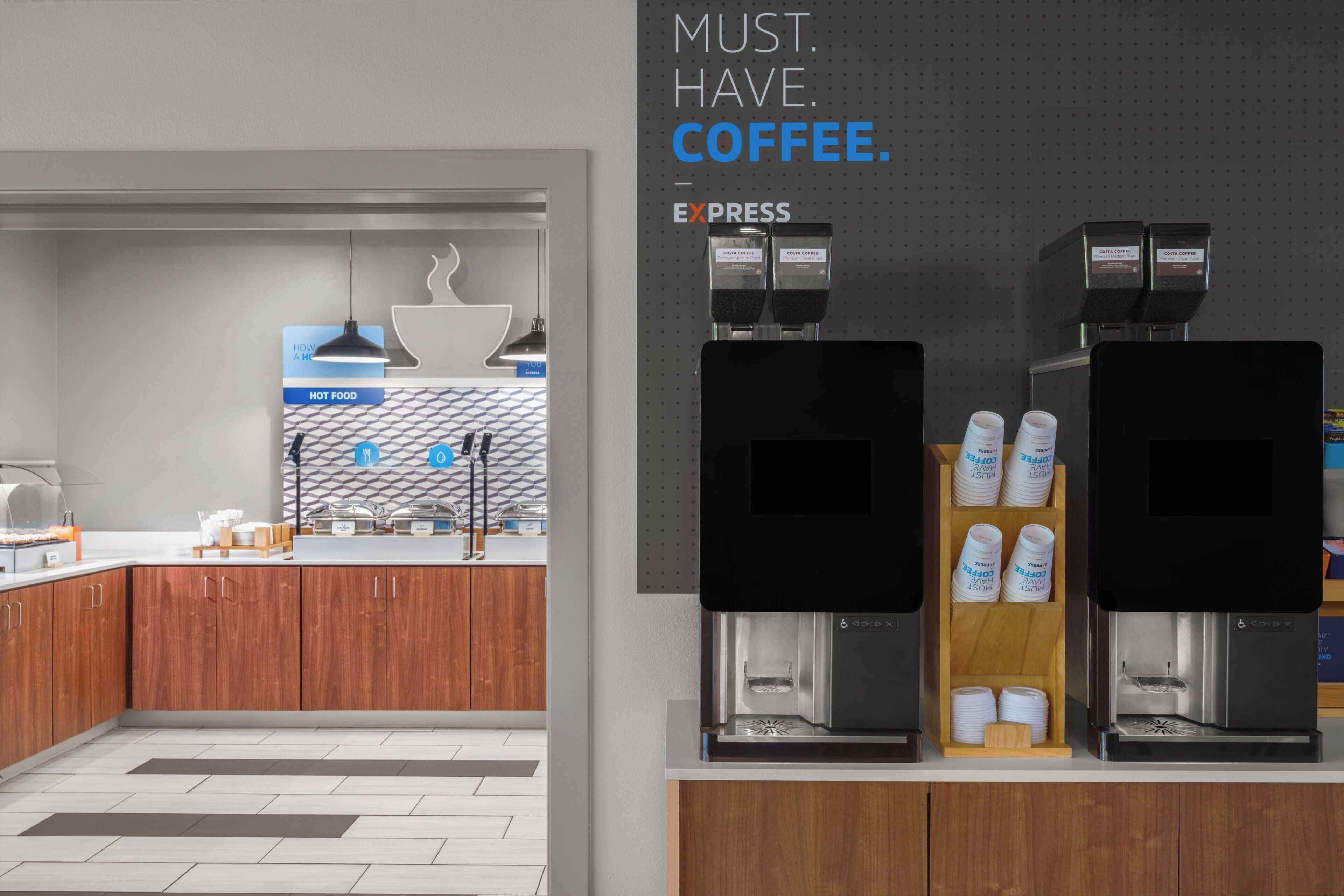 Una estación de café moderna con dos máquinas de espresso, un dispensador de Costa Coffee y un cartel que dice 'MUST. HAVE. COFFEE'.