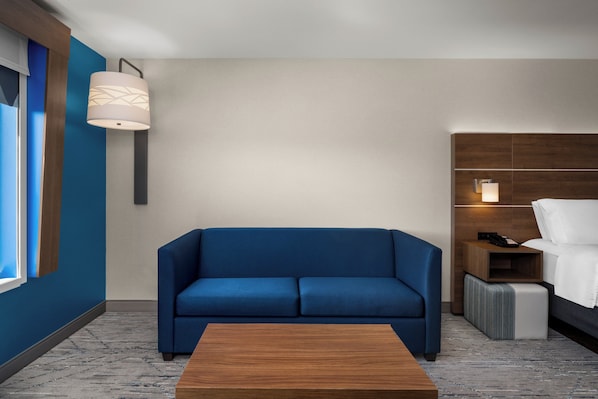 A modern hotel room with a blue sofa, a wooden coffee table, a bedside table with a lamp, and a bed with white linens. - Holiday Inn Express & Suites Salem North - Keizer by IHG (Keizer)