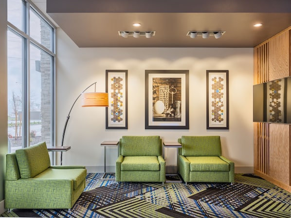 A modern hotel lobby with green armchairs, a patterned carpet, and framed artwork on the wall.