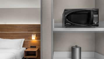 Una habitación de hotel con una cama, una mesita de noche, un microondas en un estante y un pequeño cubo de basura.