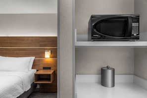 A hotel room with a bed, a nightstand, a microwave on a shelf, and a small trash can.