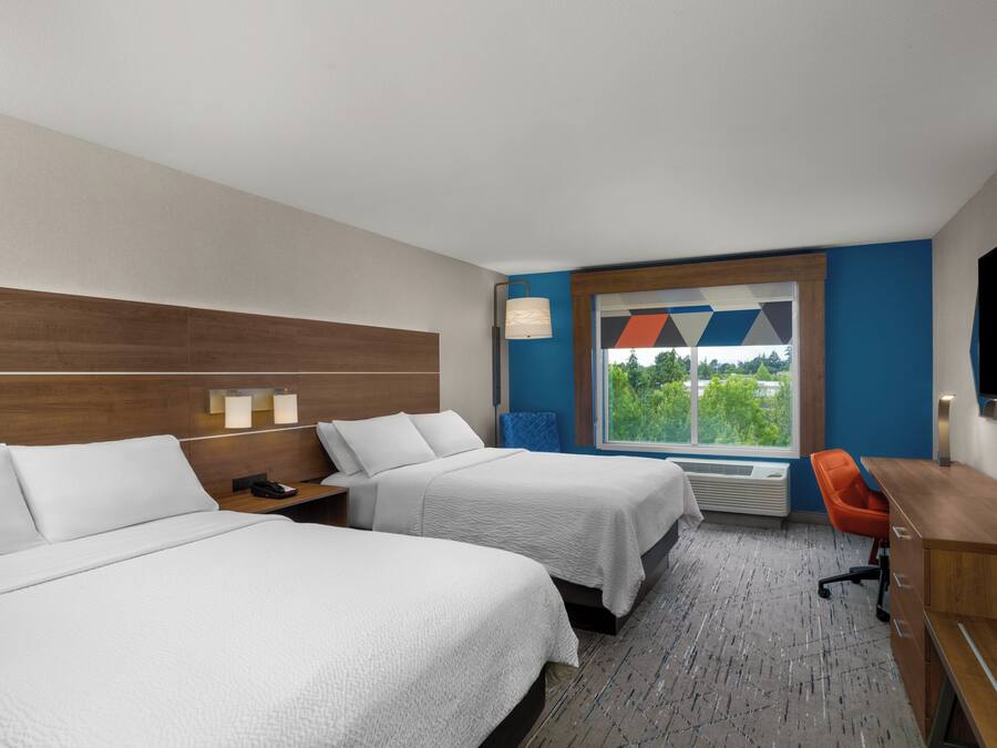 A hotel room with two beds, a desk, a chair, a TV, and a window with a view of trees.