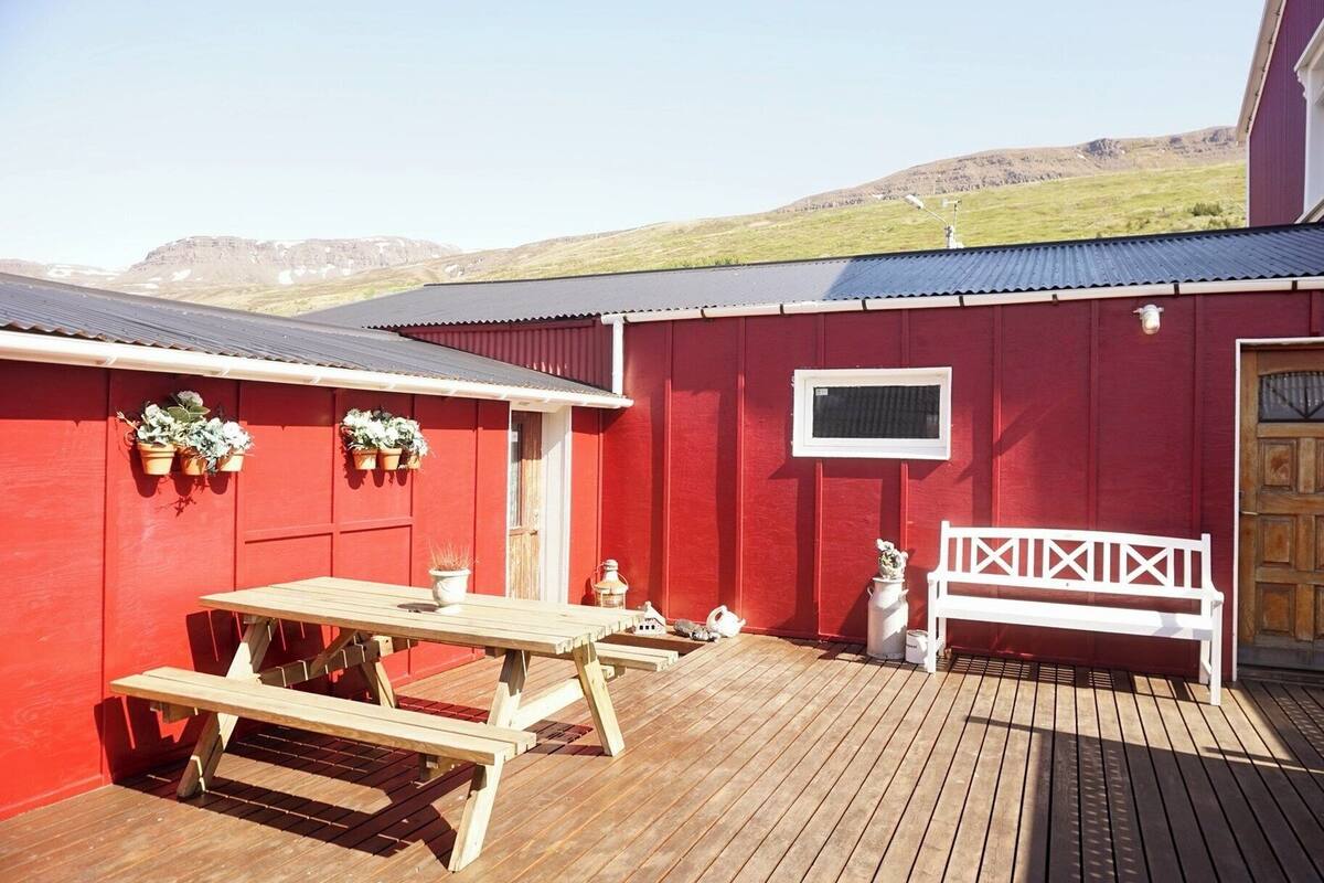 A red barn with a wooden deck, a picnic table, a bench, and potted plants.
