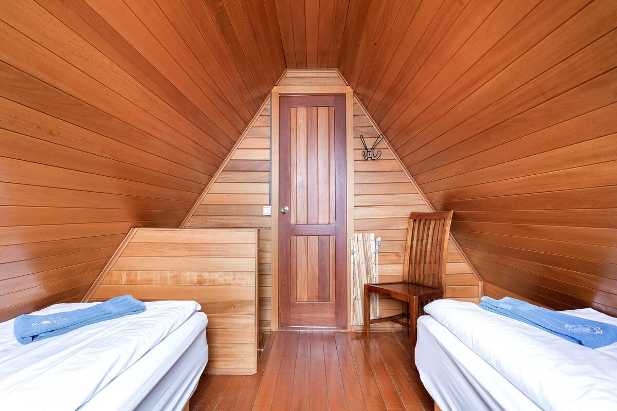 A triangular attic bedroom with wooden walls and a wooden door. There are two beds with white bedding and blue towels. A wooden chair is placed against the wall.