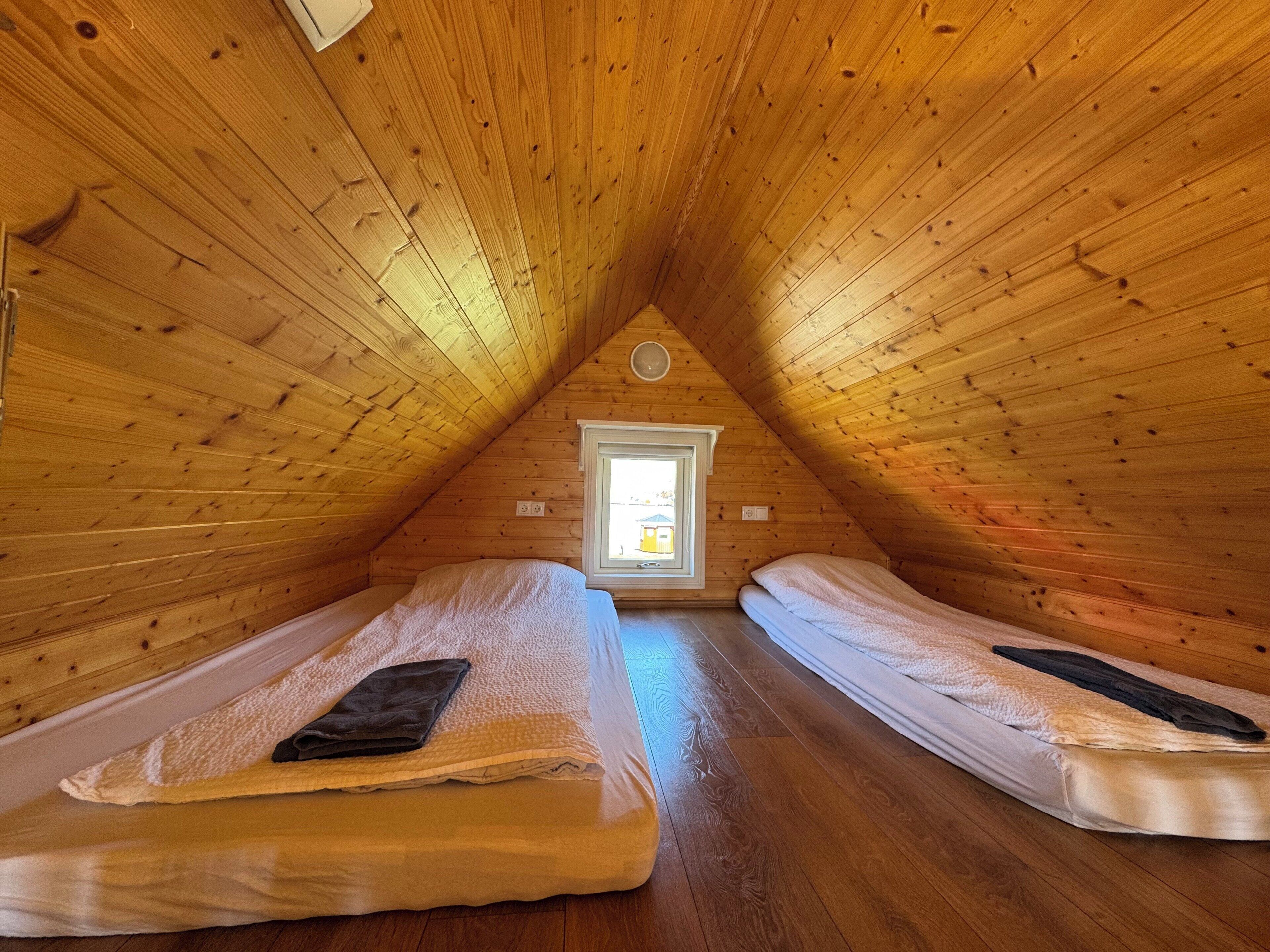 A cozy attic bedroom with wooden walls and sloped ceilings, featuring two single beds and a small window.