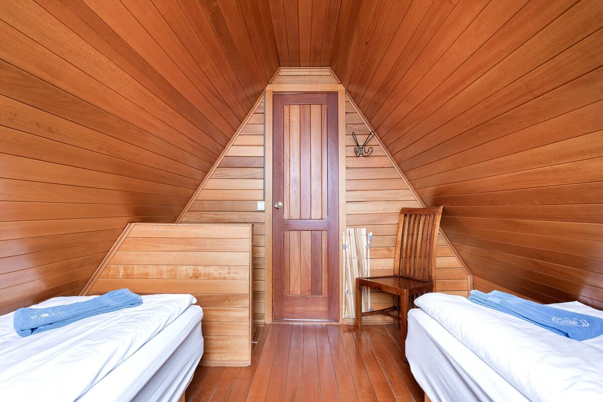 Un dormitorio en el ático con forma triangular, paredes y puerta de madera. Hay dos camas con ropa de cama blanca y toallas azules. Una silla de madera está apoyada contra la pared.