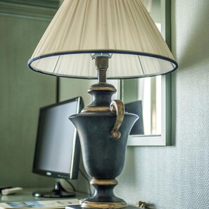 A lamp with a beige shade and a dark base, placed on a desk with a computer monitor in the background.