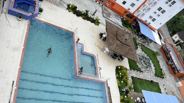 Vue aérienne d’une piscine avec un plongeoir, entourée d’un jardin et de bâtiments.