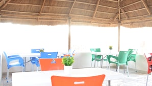 Un espace de restauration en plein air doté d’un toit de chaume, de tables blanches et de chaises en plastique aux couleurs vives.
