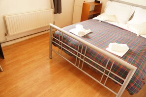 A neatly made bed with a plaid bedspread and two white towels, a wooden nightstand, and a radiator on the wall. - Beechwood House (London)